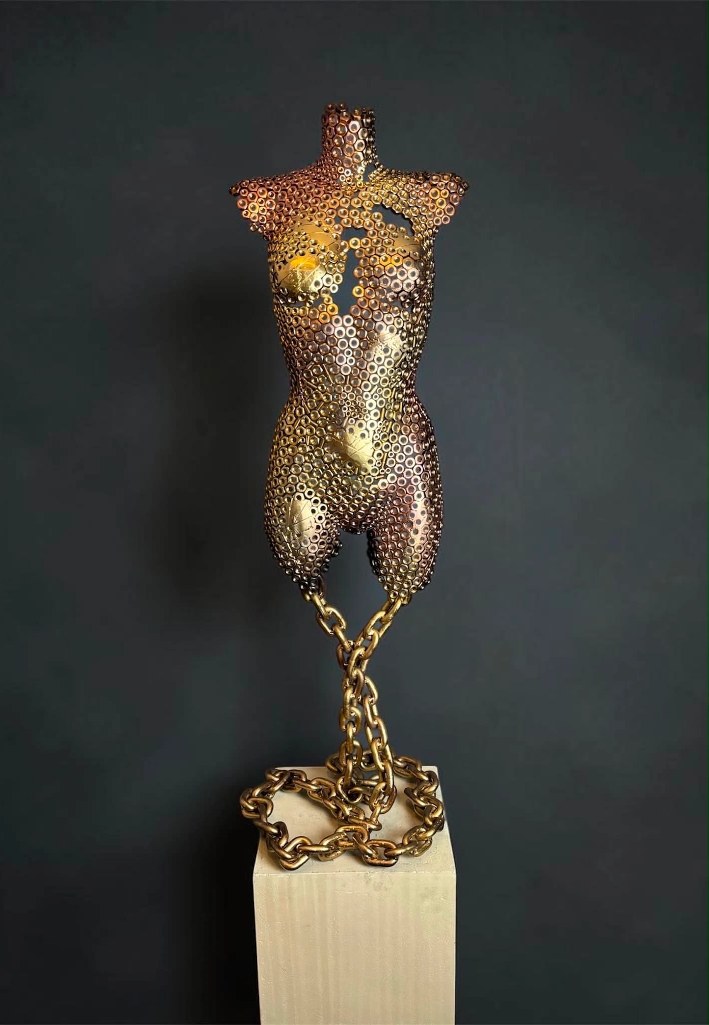 Golden Balance — goudkleurige torso van stalen moeren, hangend aan een ketting boven een betonnen sokkel